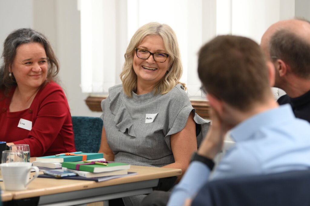 A woman in a smart grey dress, with slight ruffling around the neck and arms, smiles at a man who's back is to the camera. She is seated next to another woman in a red jumper who is slightly cropped from the photo.