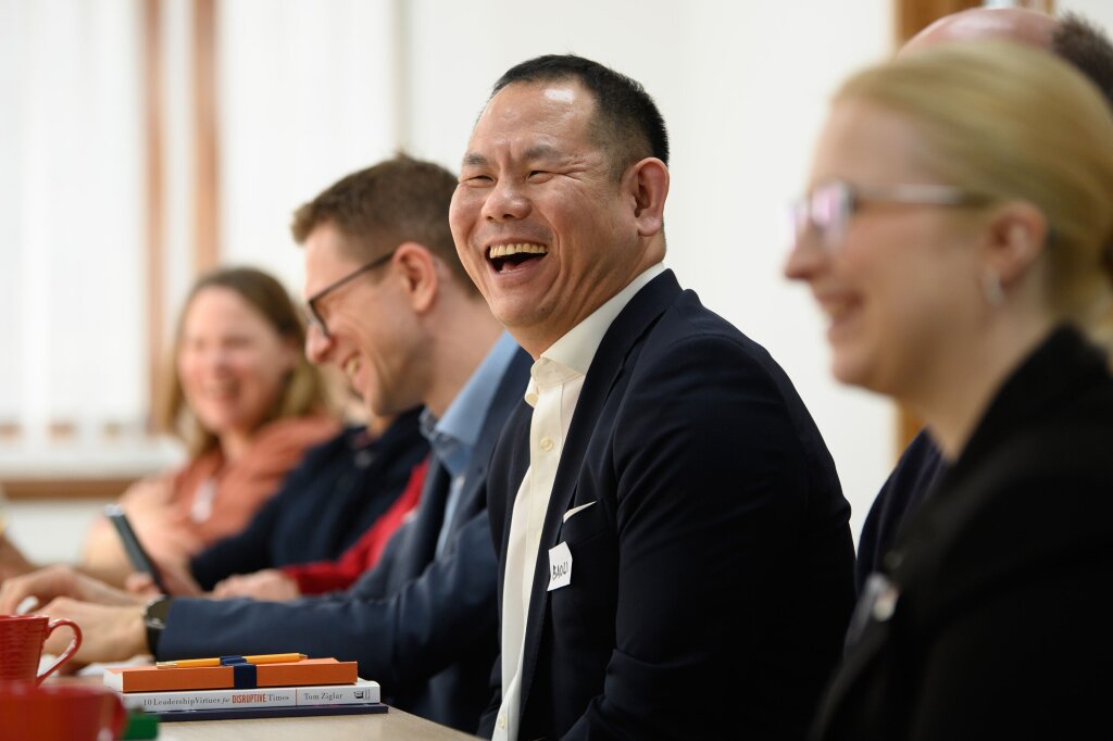A man in a white shirt and black blazer laughs. The other people sitting around the table laugh too, but they are out of focus.