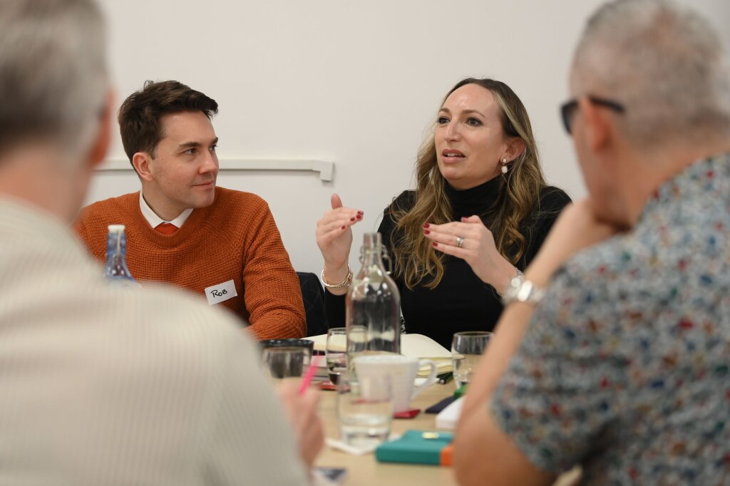 A man and a woman sit next to each other at a table covered in notebooks, coffee cups and glass water bottles. The woman is speaking, using her hands, and the man next to her is turned towards her.