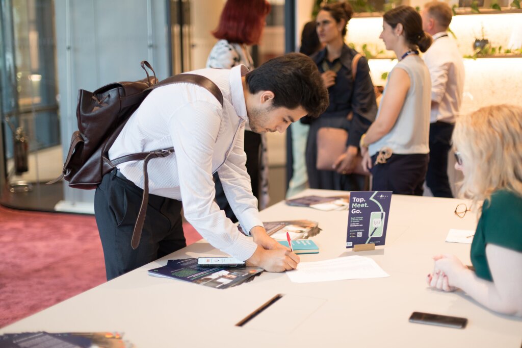 A man dressed in a suit and smart trousers leans forward on a white desk to sign a piece of paper. He is wearing a leather backpack, and it's clear from the people in the background that he is at an event.