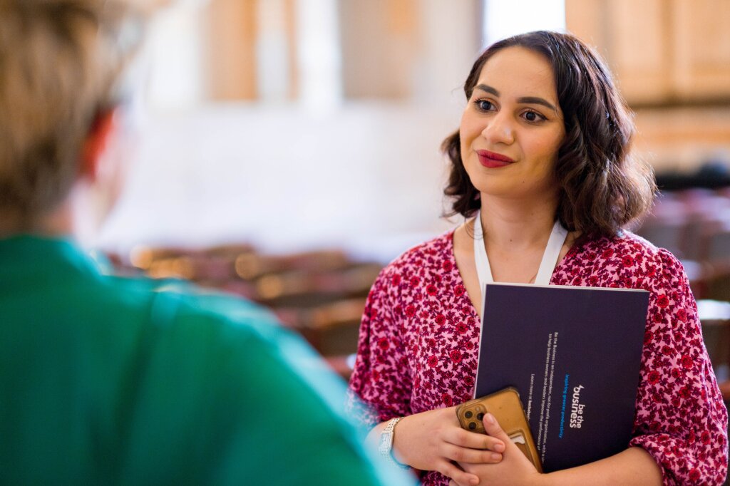 A woman with short brown hair and a pink floral blouse holds a copy of the G7 PBI to her chest. She is listening to a woman who has her back to the camera and is shown slightly out of focus.