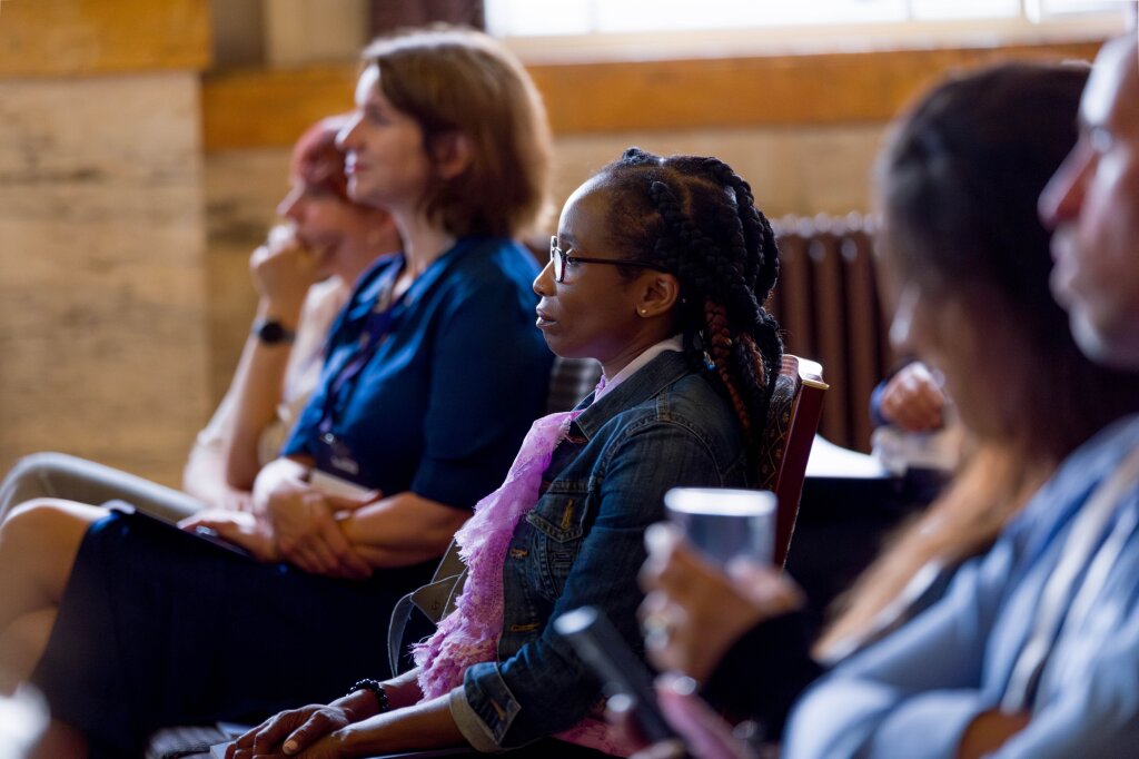 An audience listening to a speaker. The focus is on a woman with glasses and a pink scarf.