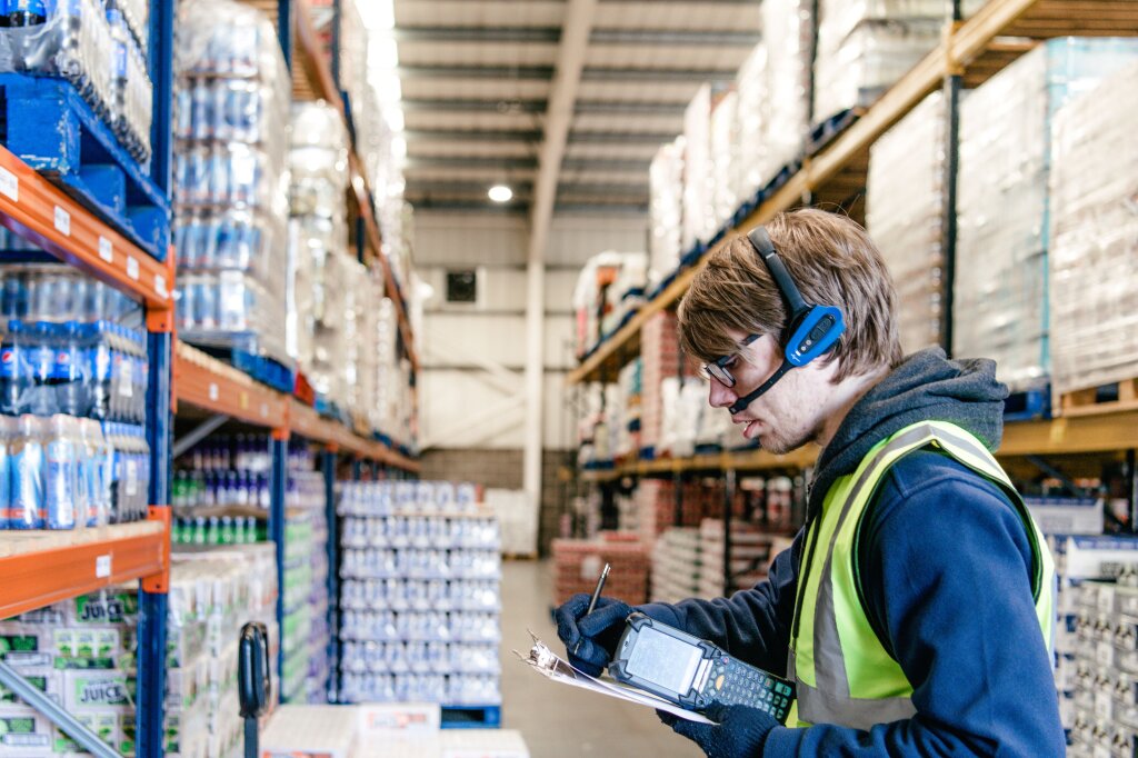 A man in a hi-viz jacket and a headset works on a clipboard in a warehouse.
