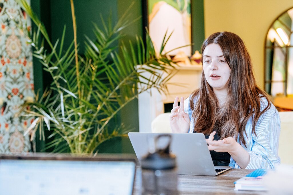 A young woman with long brown hair works on a laptop in a colourful office space. She appears to be in an online call, as she is speaking at the screen and using her hands to demonstrate her point.
