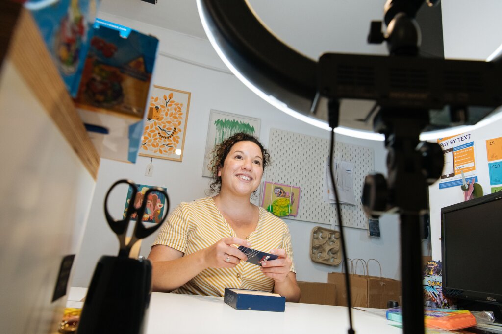 A woman dressed in a yellow and white striped blouse records herself doing a toy demonstration. She is in a brightly-lit shop, surrounded by wall art and other toys.